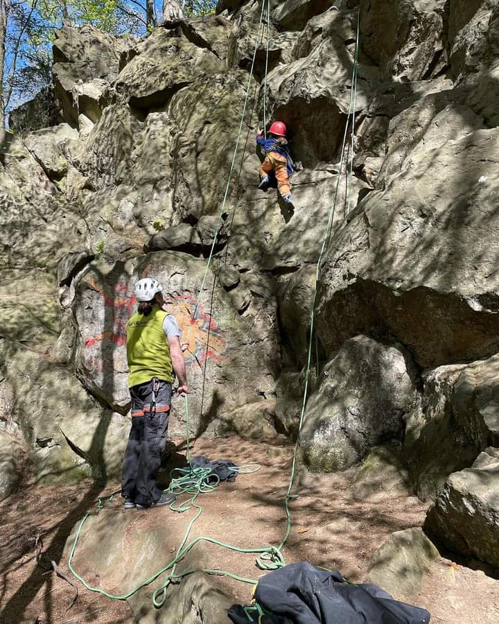 Ein Kind klettert einen Felsen hoch und wird dabei von einer Kletterteamerin gesichert.